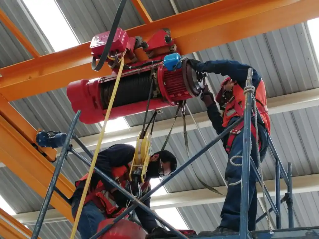 Técnicos realizando la reparación de un polipasto en altura, utilizando equipo de seguridad, mientras realizan ajustes en el motor y los componentes del sistema de elevación dentro de un entorno industrial.